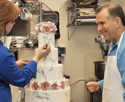 Two people decorate a multi-tiered cake with pink floral details in a kitchen setting.