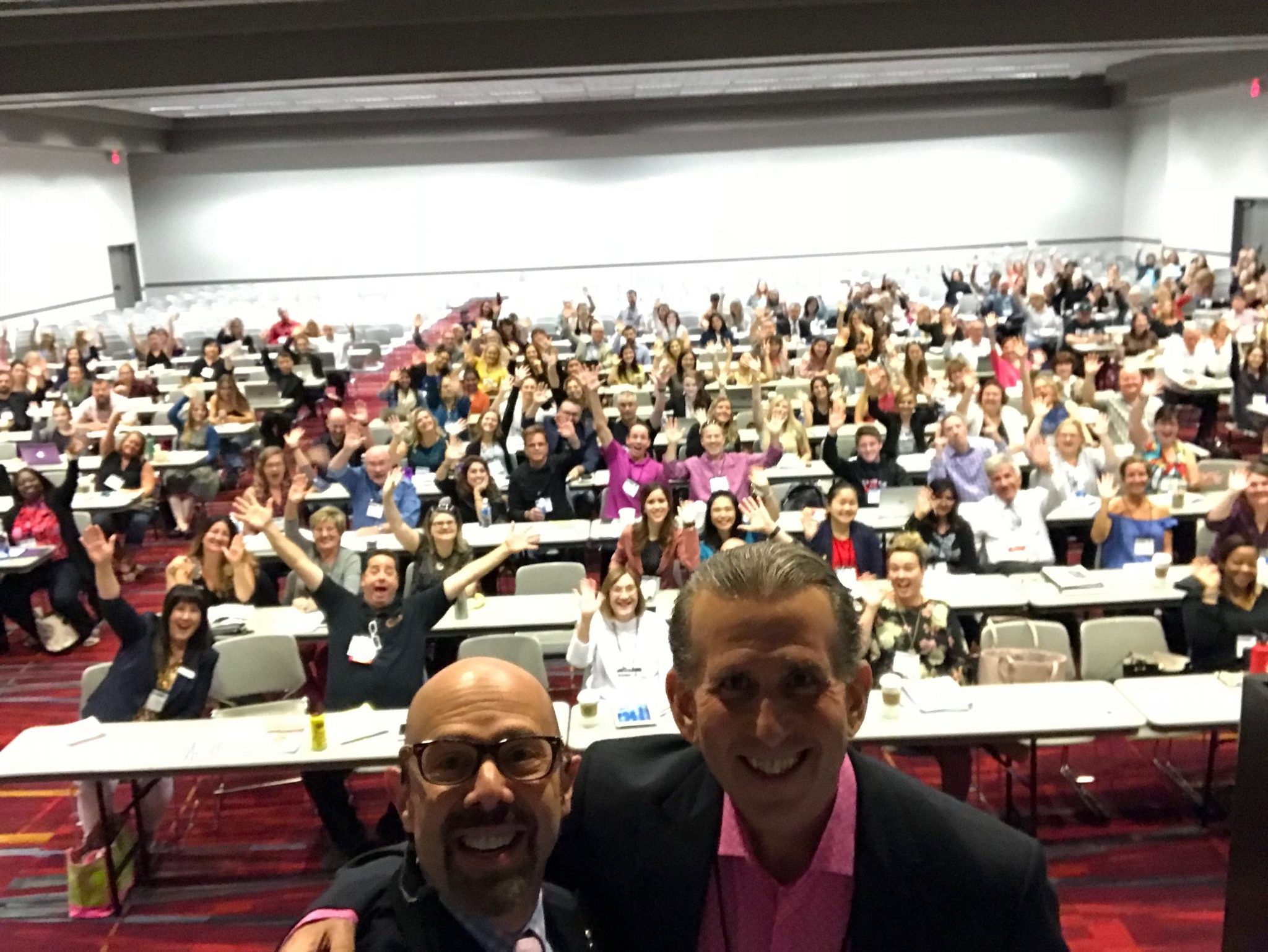 Two people take a selfie in front of a large group seated in a conference room. The audience waves and smiles.