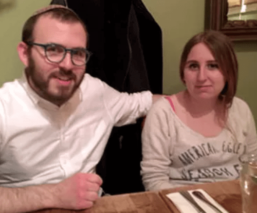 A man and a woman sit together at a table in a restaurant. The man wears glasses and a white shirt, and the woman wears a light-colored top. A coat hangs in the background.