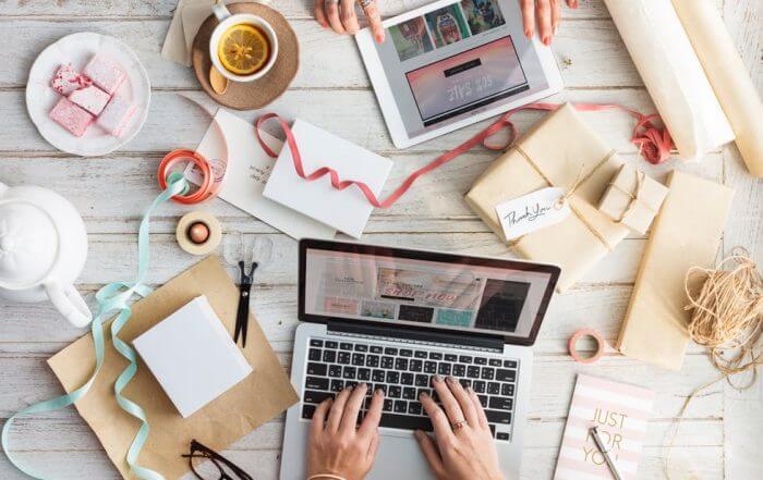 Hands using a laptop and tablet on a table with gift wrapping materials, ribbon, cards, tea, and sweets.