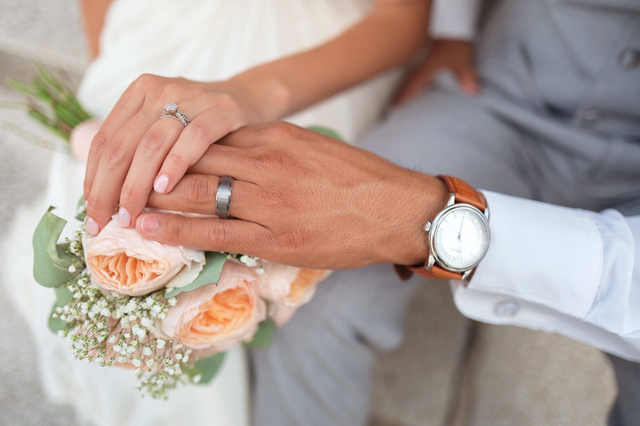 Close-up of a couple holding hands over a bouquet of pink roses. The woman has a diamond ring and the man wears a watch and wedding band.