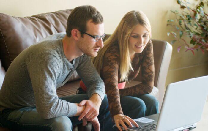 A man and woman sit on a couch, focused on a laptop screen, with the woman pointing at the display.