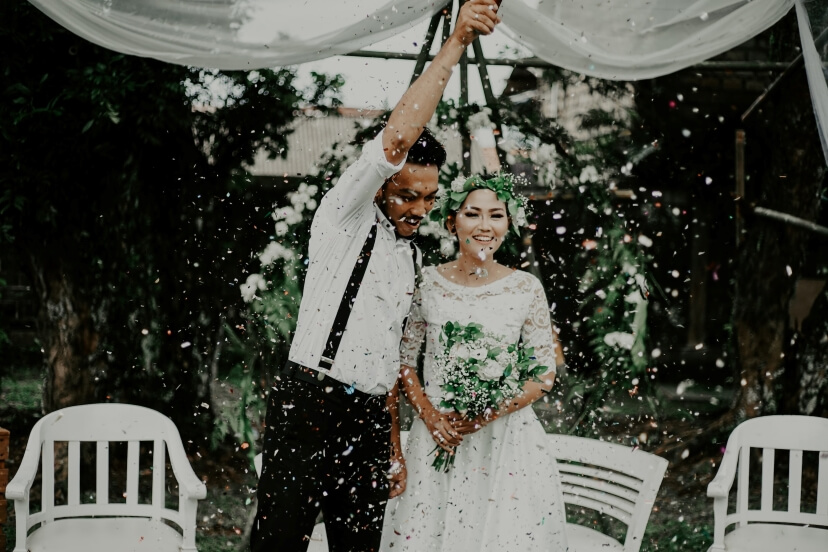 A couple celebrates under a canopy with confetti falling, surrounded by chairs and greenery.