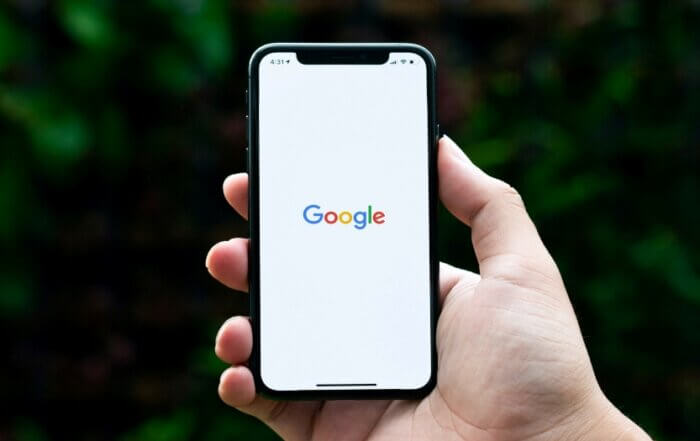 Hand holding a smartphone displaying the Google logo on the screen against a blurred green background.