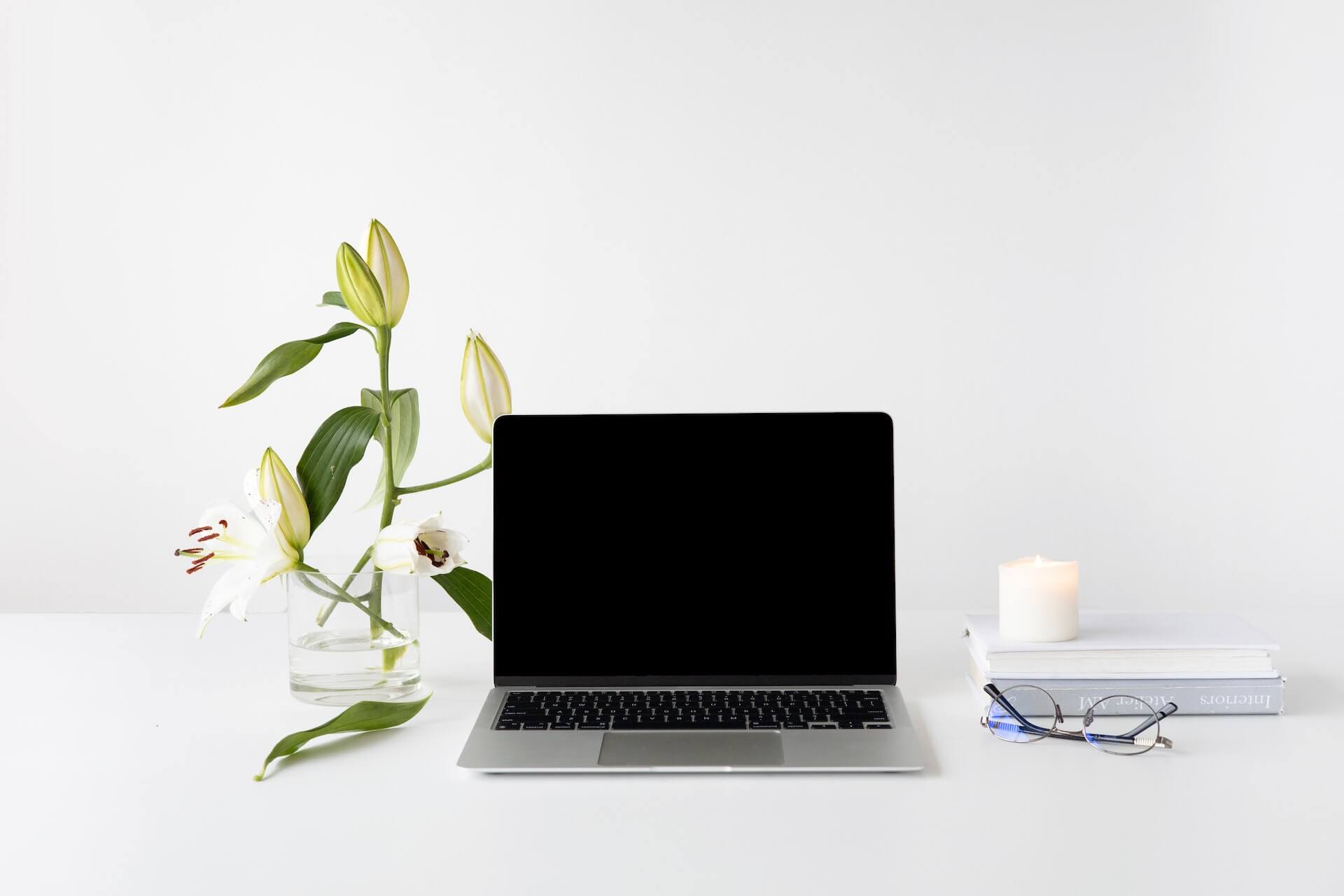 Laptop on a white desk with a blank screen. Vase with lilies, a candle, a stack of books, and glasses are also on the desk.
