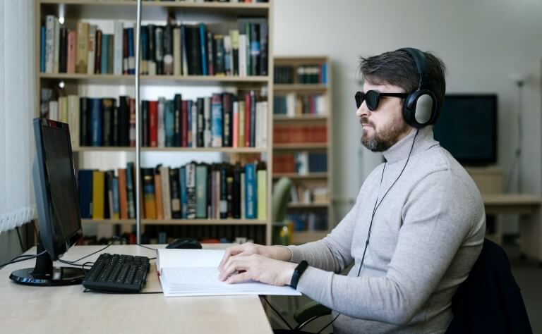 A man wearing sunglasses and headphones uses a computer with braille materials in front of bookshelves.