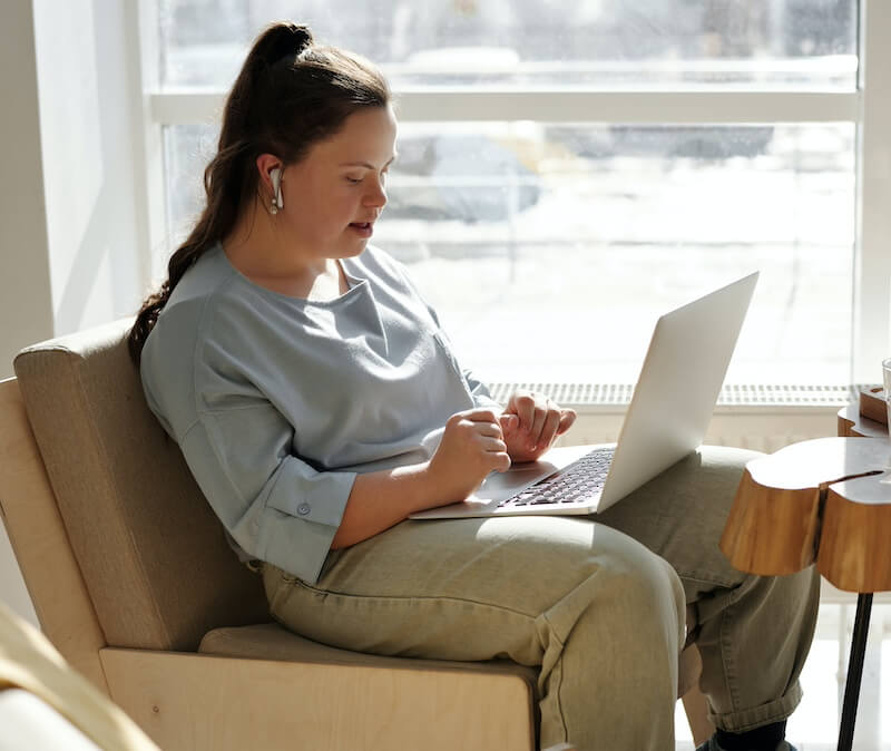 A young women visiting an ADA compliant website A person sits on a chair using a laptop, wearing earbuds and a casual blue shirt with light green pants, near a sunlit window.